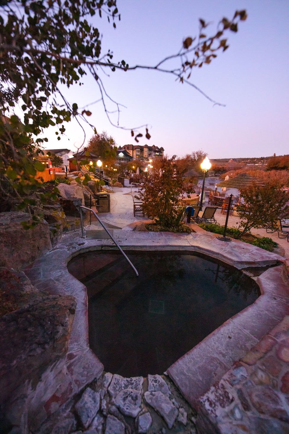 A dark, stone-lined hot tub, with a metal railing dipping into the water, is nestled among rocks and trees at dusk, overlooking an illuminated resort area.