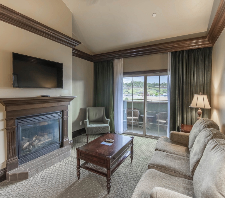 A living room features a fireplace with TV above, sofa, and coffee table, overlooking a balcony with a scenic view, adorned with decorative wooden trim.