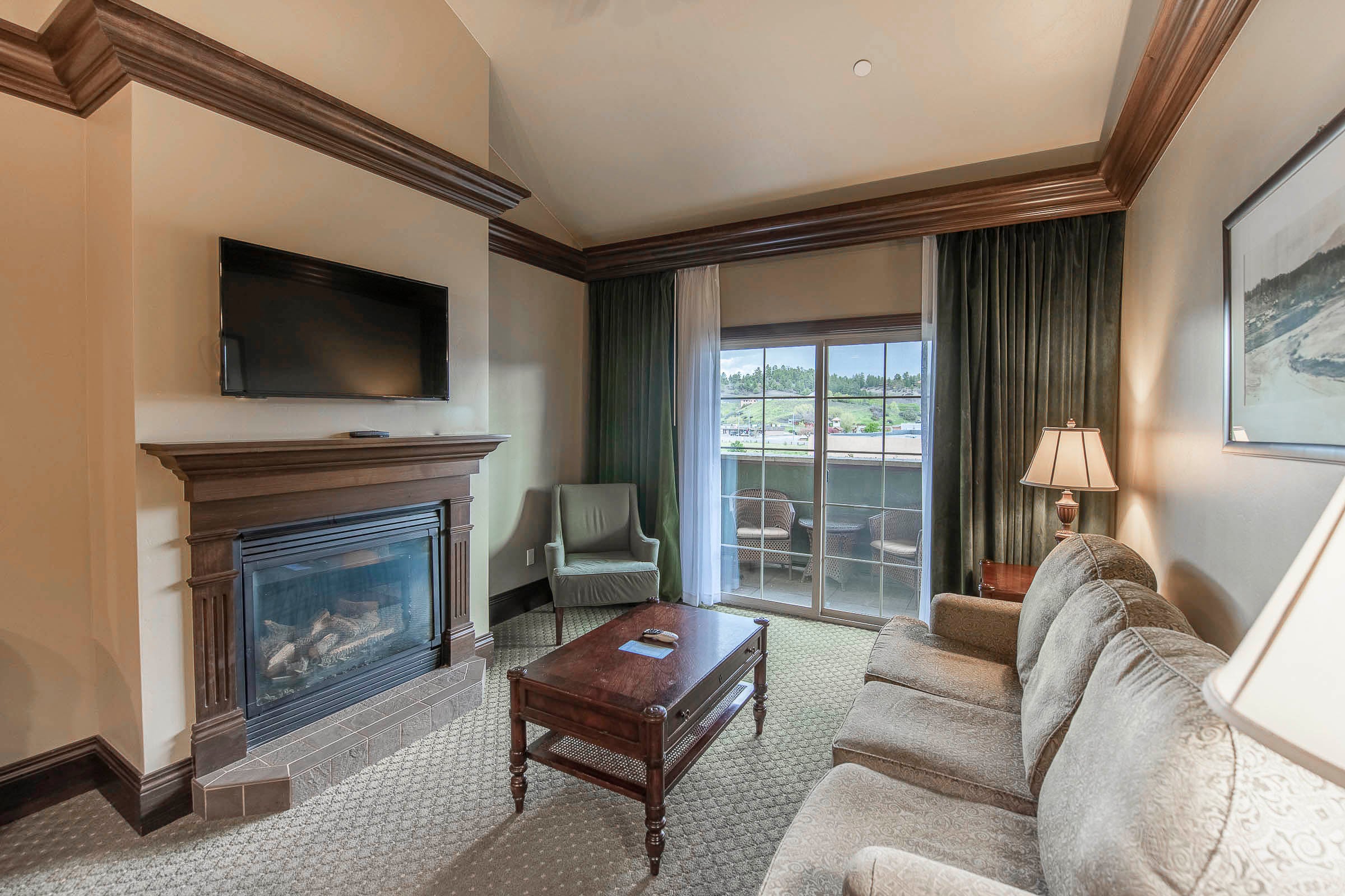 A living room features a fireplace with TV above, sofa, and coffee table, overlooking a balcony with a scenic view, adorned with decorative wooden trim.