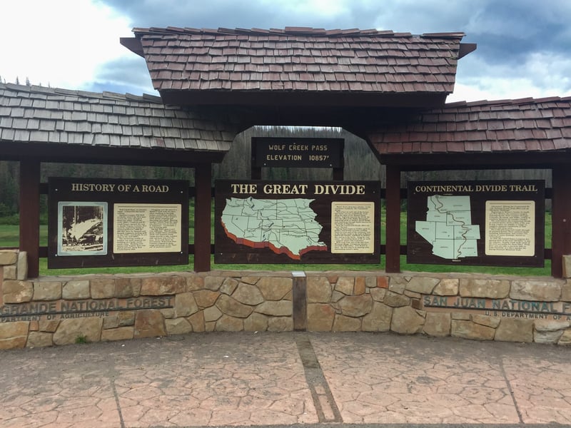 A large brown multi-panel sign with a shingled roof presents information on a stone wall, backed by a forest and cloudy sky.
WOLF CREEK PASS ELEVATION 10857' HISTORY OF A ROAD THE GREAT DIVIDE CONTINENTAL DIVIDE TRAIL GRANDE NATIONAL FOREST DEPARTMENT OF AGRICULTURE SAN JUAN NATIONAL FOREST U.S. DEPARTMENT OF AGRICULTURE