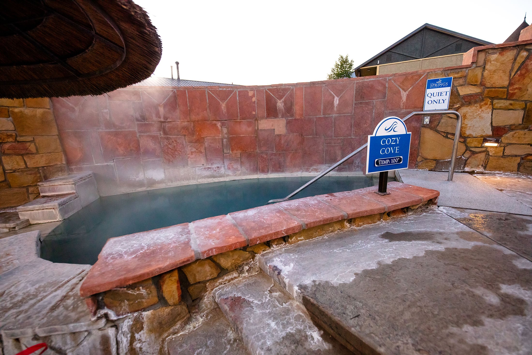 A hot spring pool steams, set into irregular reddish stone walls and wet concrete steps.
Text: THE SPRINGS QUIET ONLY, COZY COVE TEMP: 100°