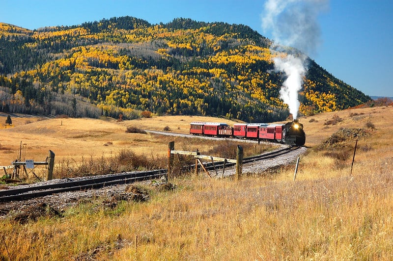 A black steam train with red cars travels on tracks through a golden field with an autumn forest mountain. Sign: PRIVATE PROPERTY POSTED.
