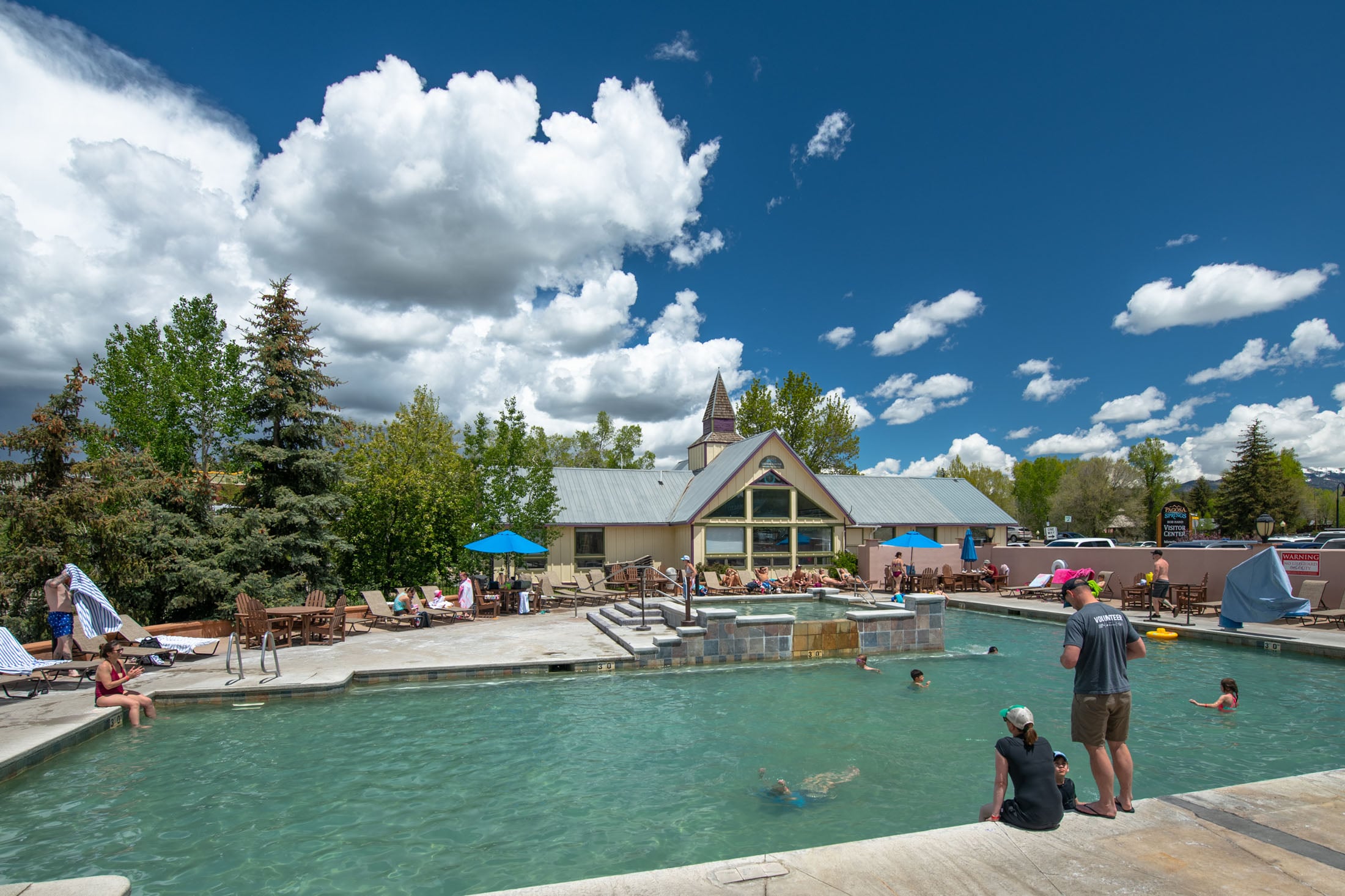 People swim and lounge in a large outdoor pool area on a sunny day. A building with a peaked roof and trees line the background. Text: THE SPRINGS RESORT & SPA, PAGOSA SPRINGS VISITOR CENTER, WARNING NO DIVING, WARNING, VOLUNTEER, 3-0.