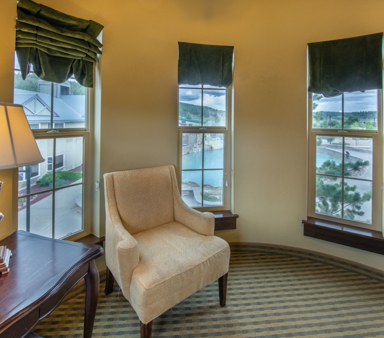A beige armchair sits in a curved room with three windows, offering views of a pool, trees, and distant buildings. A lamp rests on a dark table nearby.