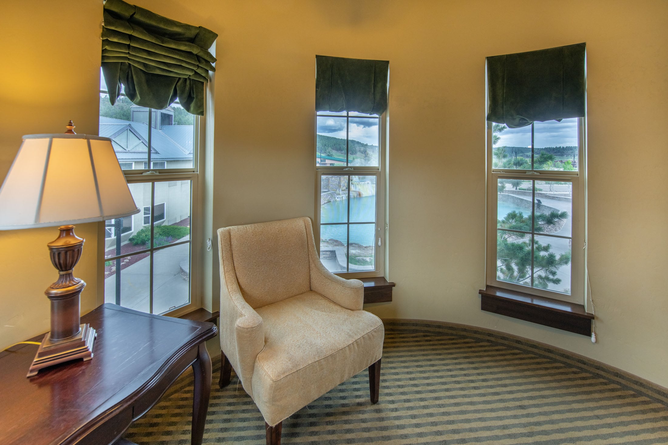 A beige armchair sits in a curved room with three windows, offering views of a pool, trees, and distant buildings. A lamp rests on a dark table nearby.