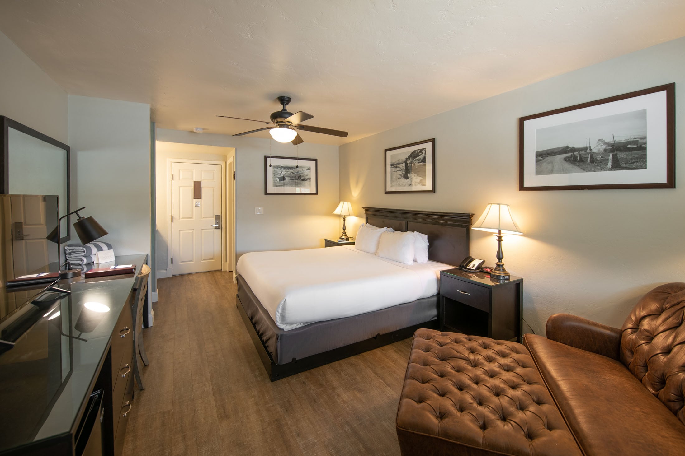 A white-linened queen bed occupies a furnished hotel room with a dark desk, leather armchair, nightstands with lamps, ceiling fan, and wooden flooring.