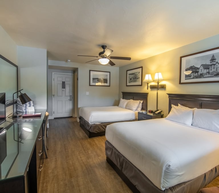 Two queen beds stand in a well-lit hotel room with light green walls and wood flooring. A desk with TV, coffee maker, and ceiling fan furnish the space, complemented by framed artwork.