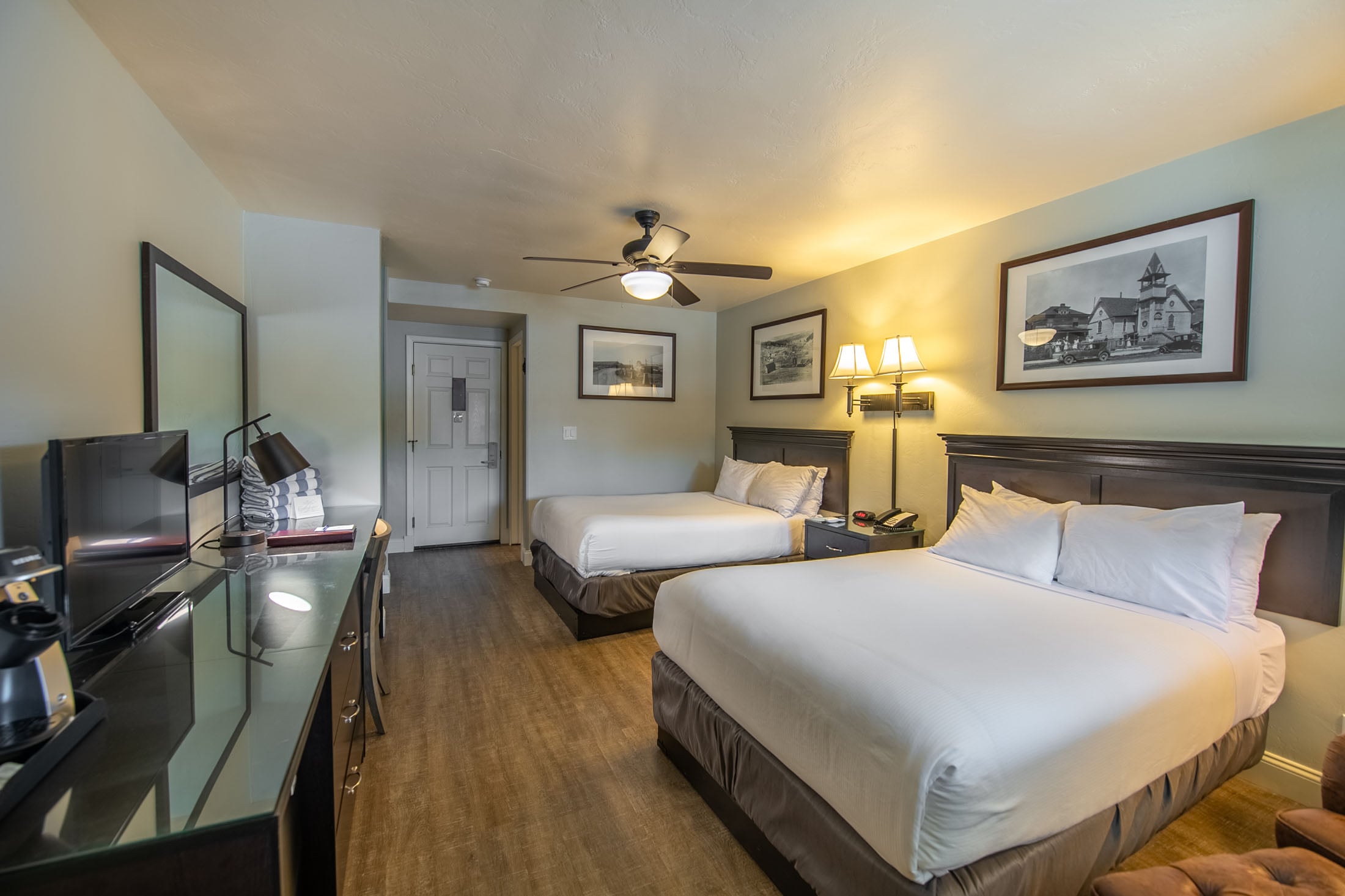 Two queen beds stand in a well-lit hotel room with light green walls and wood flooring. A desk with TV, coffee maker, and ceiling fan furnish the space, complemented by framed artwork.