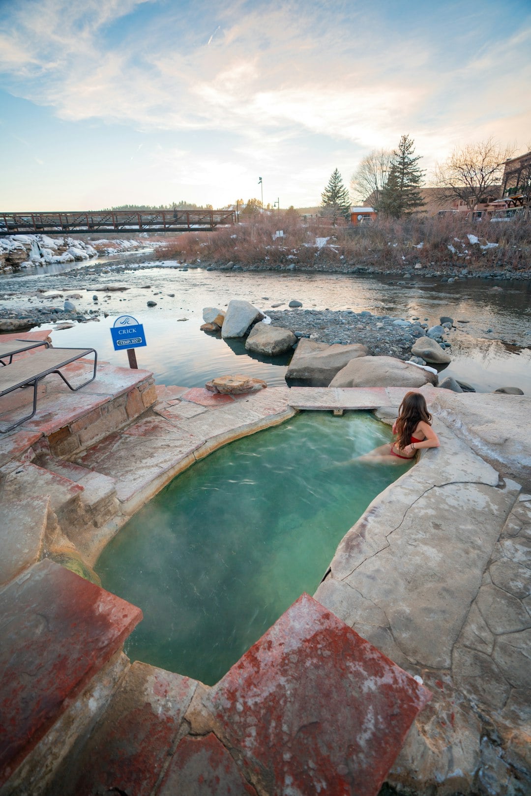 Woman in red bikini bathes in a steaming green hot spring beside a rocky river, with a bridge in the distance under a cloudy sky. Sign: CRICK TEMP. 100°