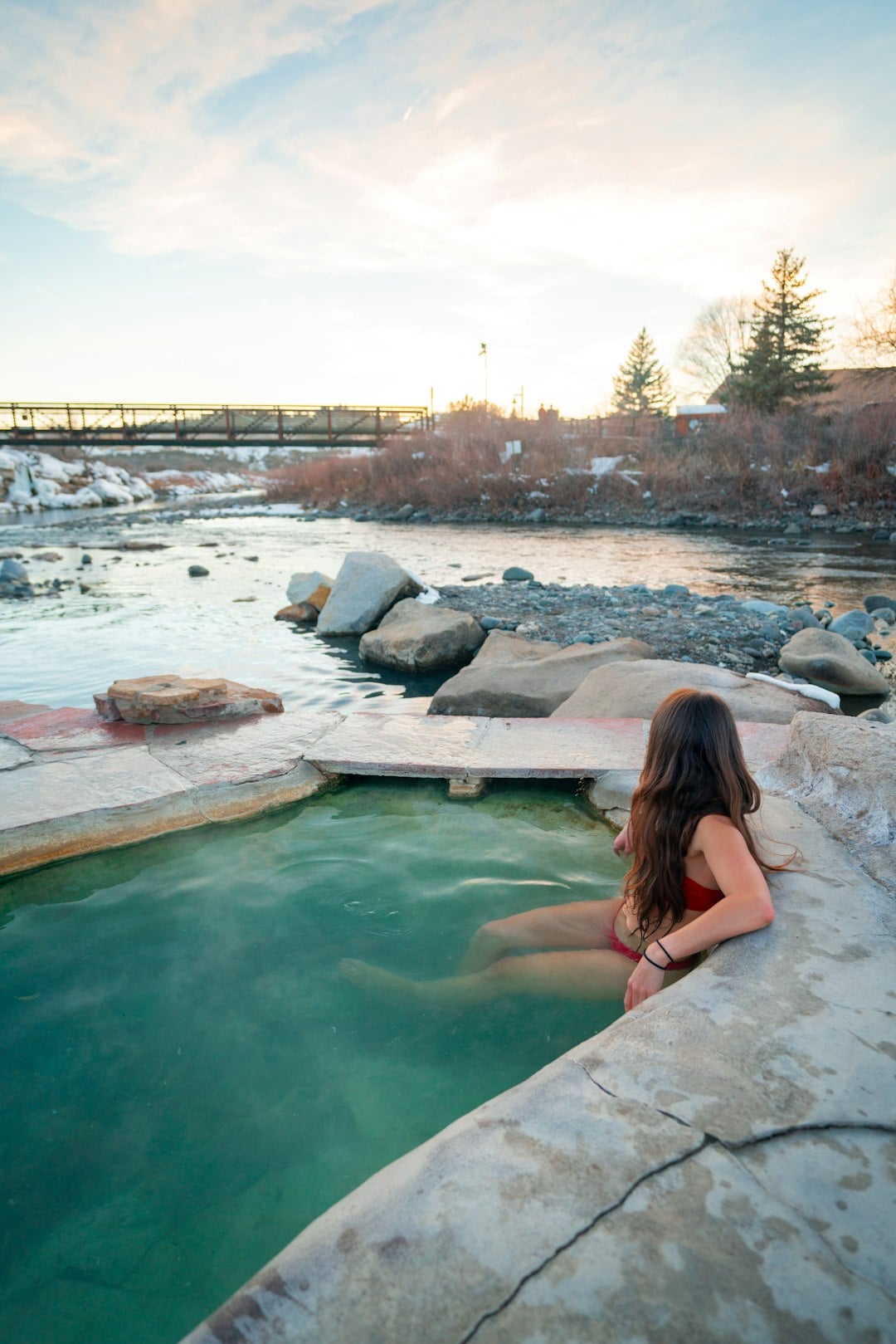 A woman in a red bikini relaxes in a steaming natural hot spring pool next to a rocky river, with a distant bridge and snowy banks under a sunset sky.