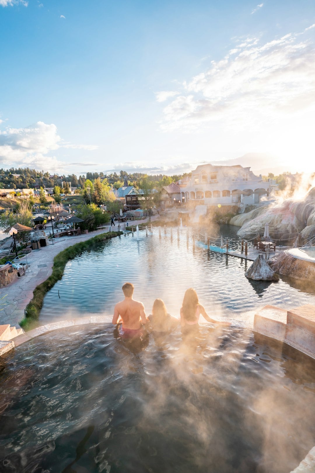 Three people relax in a steaming hot spring pool, overlooking a large natural pool and resort buildings under a bright, sunny sky with visible steam.