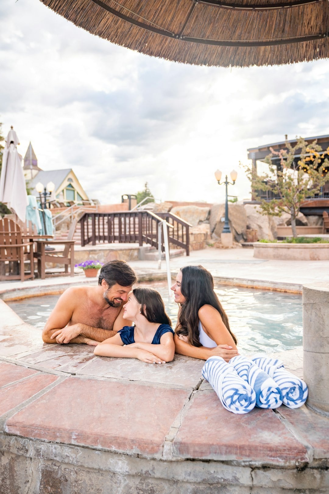 A family of three relaxes in an outdoor hot tub, smiling at each other. The resort setting includes buildings, stairs, and mountains under a bright, cloudy sky. Three striped towels are on the tub's edge.