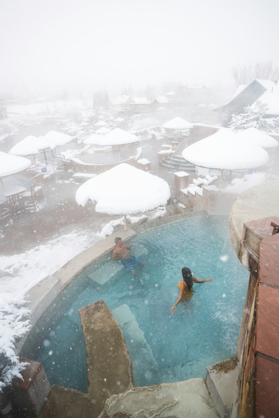 A couple relaxes in a steaming outdoor hot tub amidst heavily falling snow, which covers resort umbrellas and distant buildings.