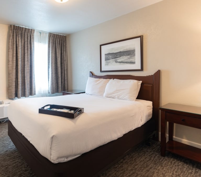 A white-linened bed with a dark headboard holds a tray of items, situated in a hotel room with beige walls, drawn curtains, and a landscape print above it.