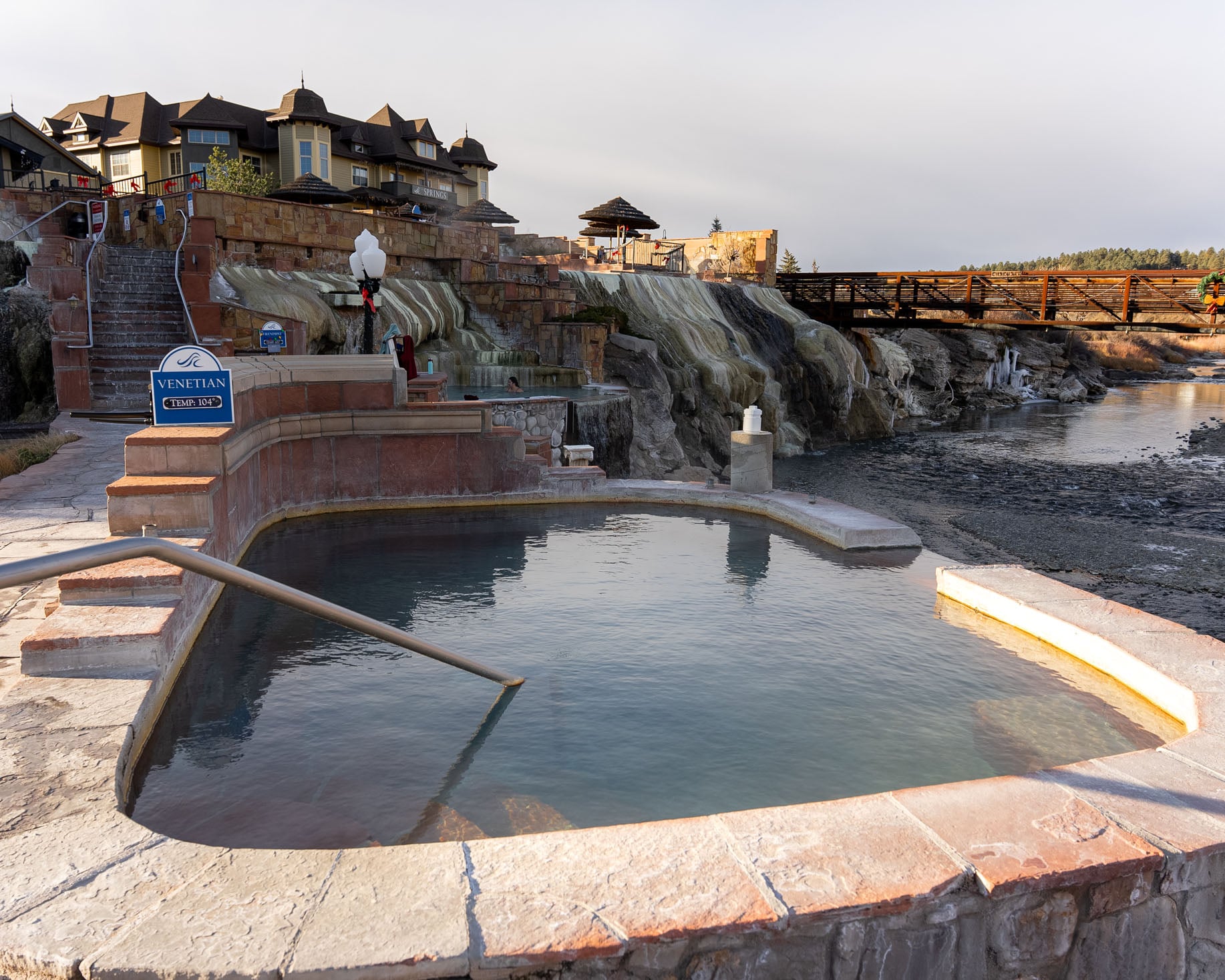 An outdoor hot spring pool holds blue-green water with a metal handrail, flanked by tiered rock waterfalls and a large resort building, with a river and bridge beyond. VENETIAN TEMP: 104°, MANDARIN TEMP: 102°, THE SPRINGS, CUMBRE & TOLTEC.