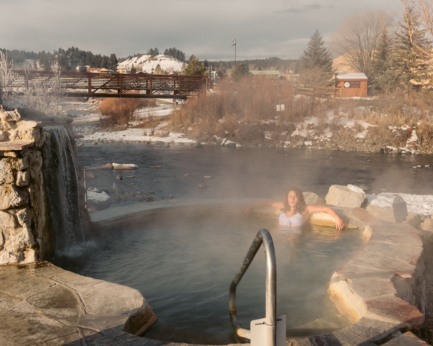 A woman relaxes in a steaming rock-lined hot spring beside a river with snowy banks, a waterfall, and a bridge; distant shack reads: WE ARE OPEN!