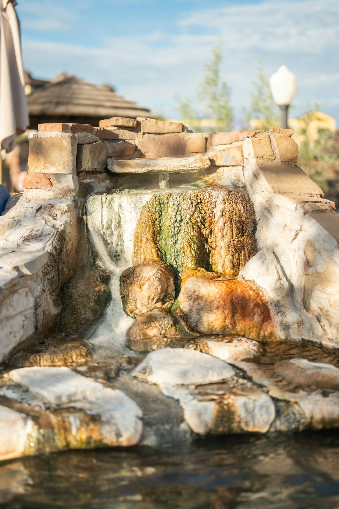 A multi-tiered rock and brick waterfall cascades water down its algae-covered, mineral-stained surfaces into a pool, set outdoors with a thatched roof building and bright sky behind it.