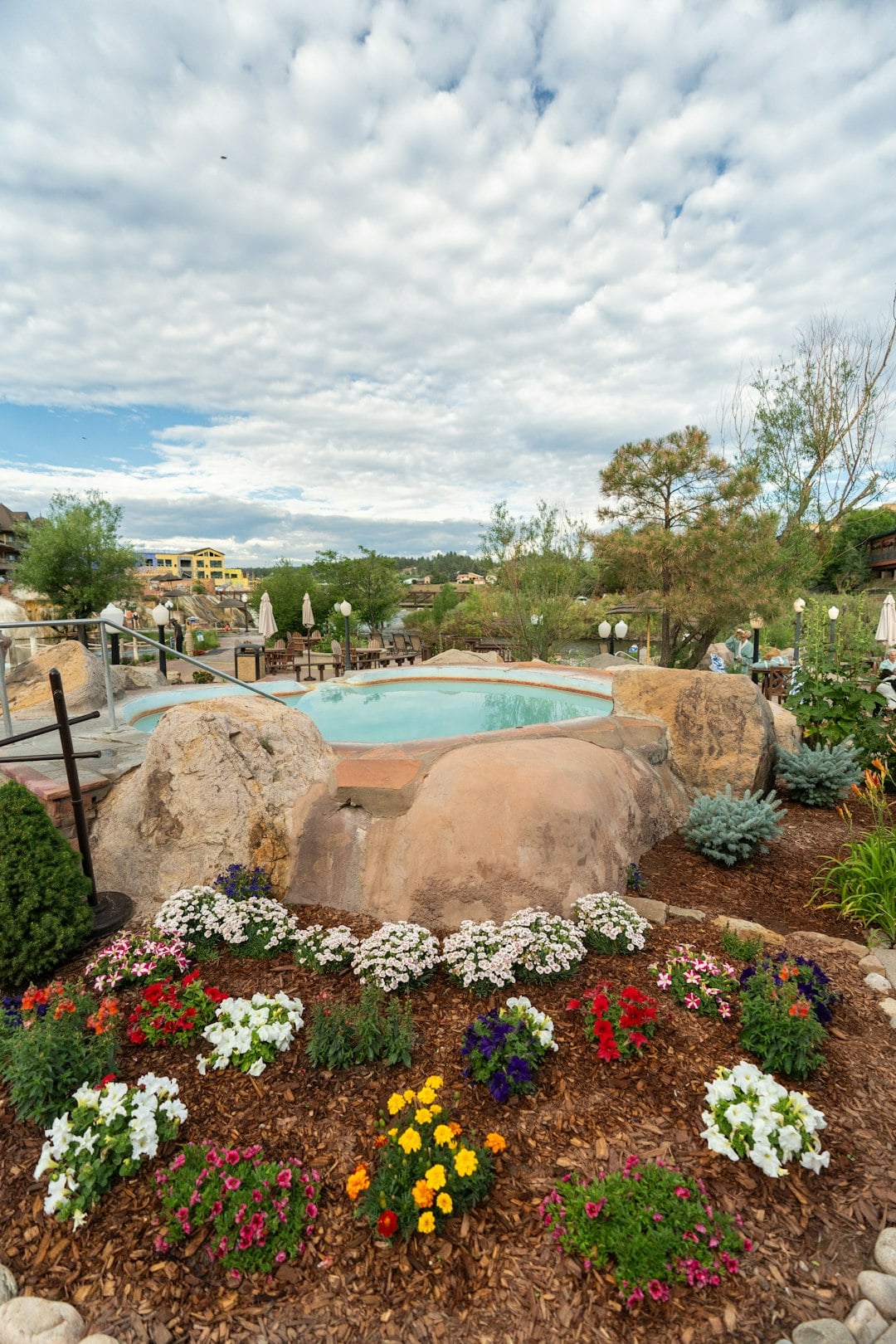 A rock-encased hot tub with light blue water is surrounded by vibrant flower beds. It's set in an outdoor resort area with distant buildings and trees under a cloudy sky.