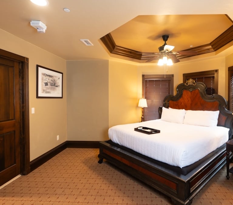 A large bed with an ornate dark wood headboard dominates a bedroom, featuring white bedding with a tray, textured brown carpet, and a tray ceiling with a fan.