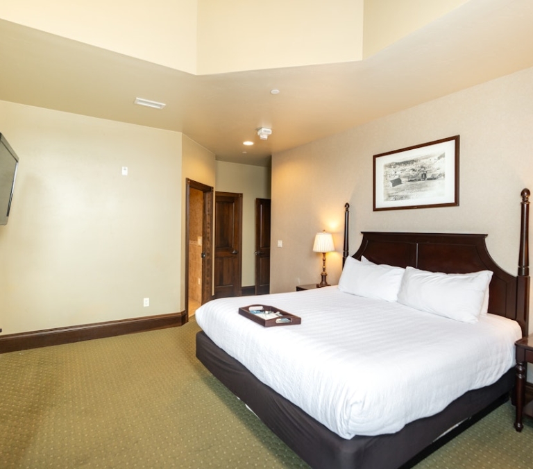 A dark wood king bed with white linens and a tray sits centrally in a spacious bedroom. Flanked by lit lamps, a TV and dresser complete the carpeted interior.