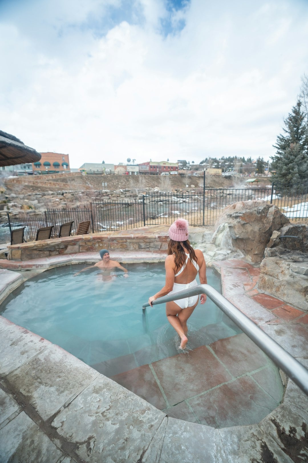 A woman in a white swimsuit and pink hat steps into a steamy outdoor hot spring, holding a rail. A man soaks nearby. Stone-edged pool, with winter foliage and a distant town under a cloudy sky.
