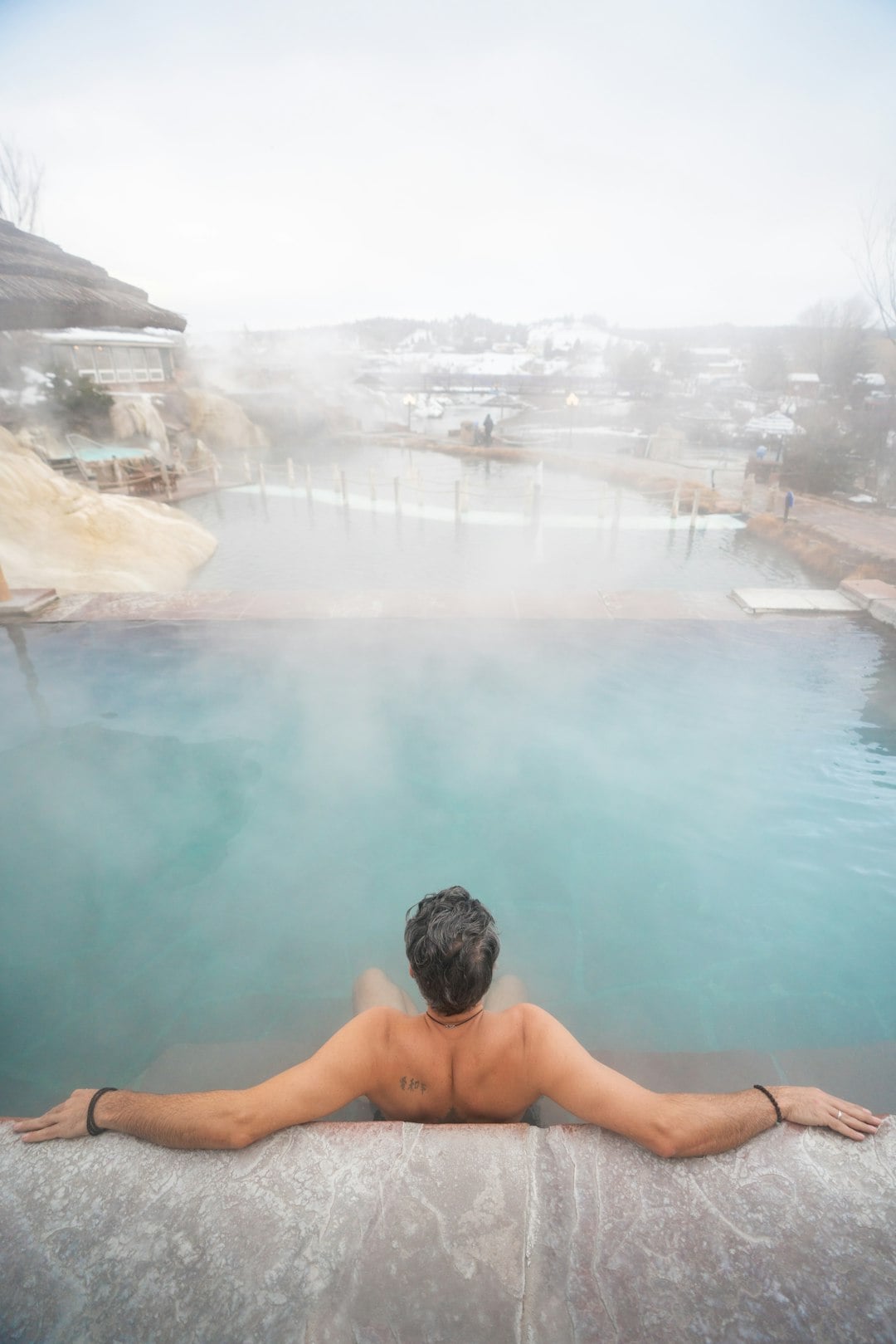 A shirtless person with grey hair relaxes in a steaming turquoise hot spring, looking over a snowy landscape with other pools and distant buildings. Back tattoo: 泰和