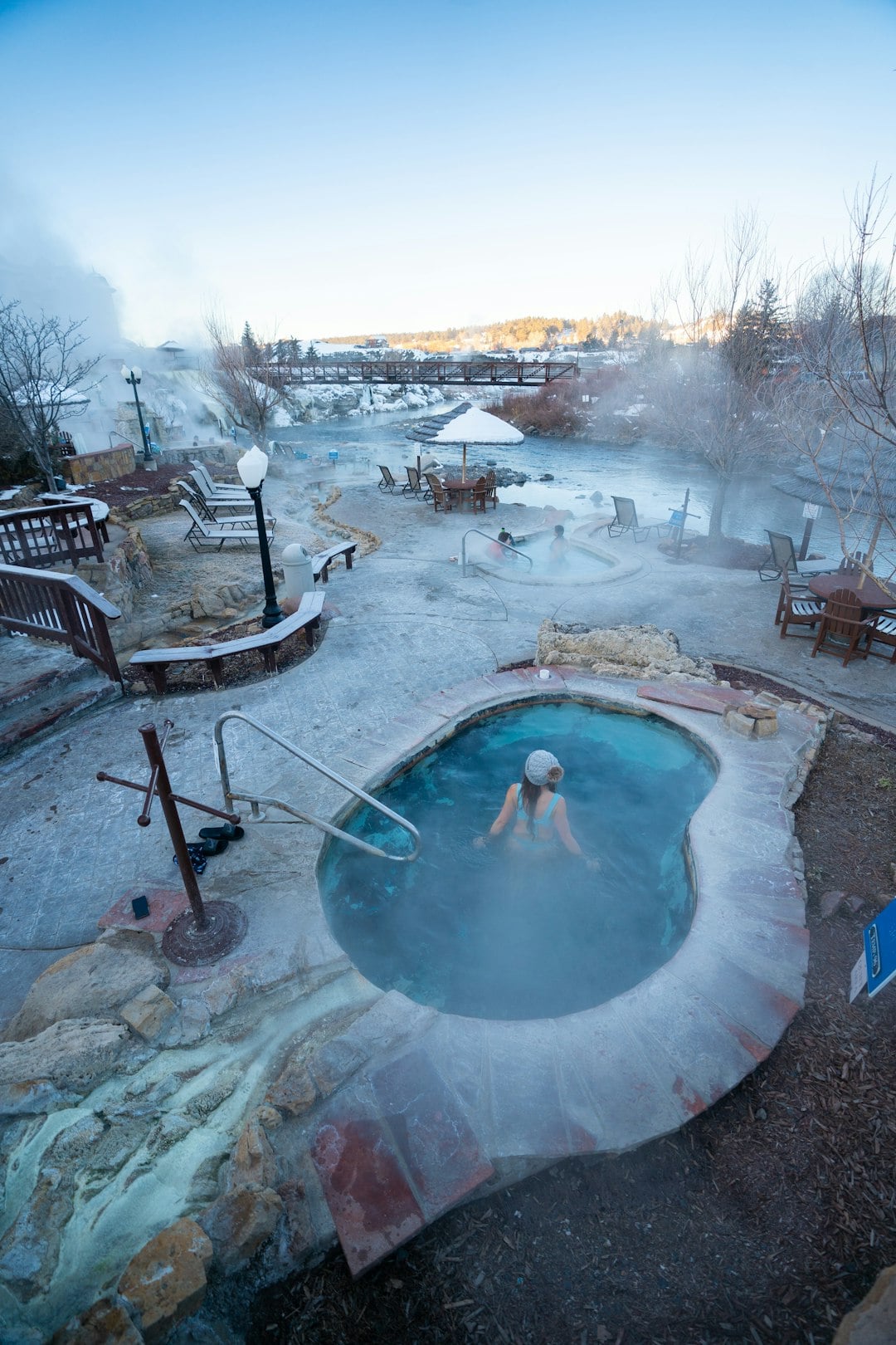 A woman relaxes in a steaming outdoor hot tub, surrounded by other hot pools and a river in a snow-covered resort area with a bridge in the distance.