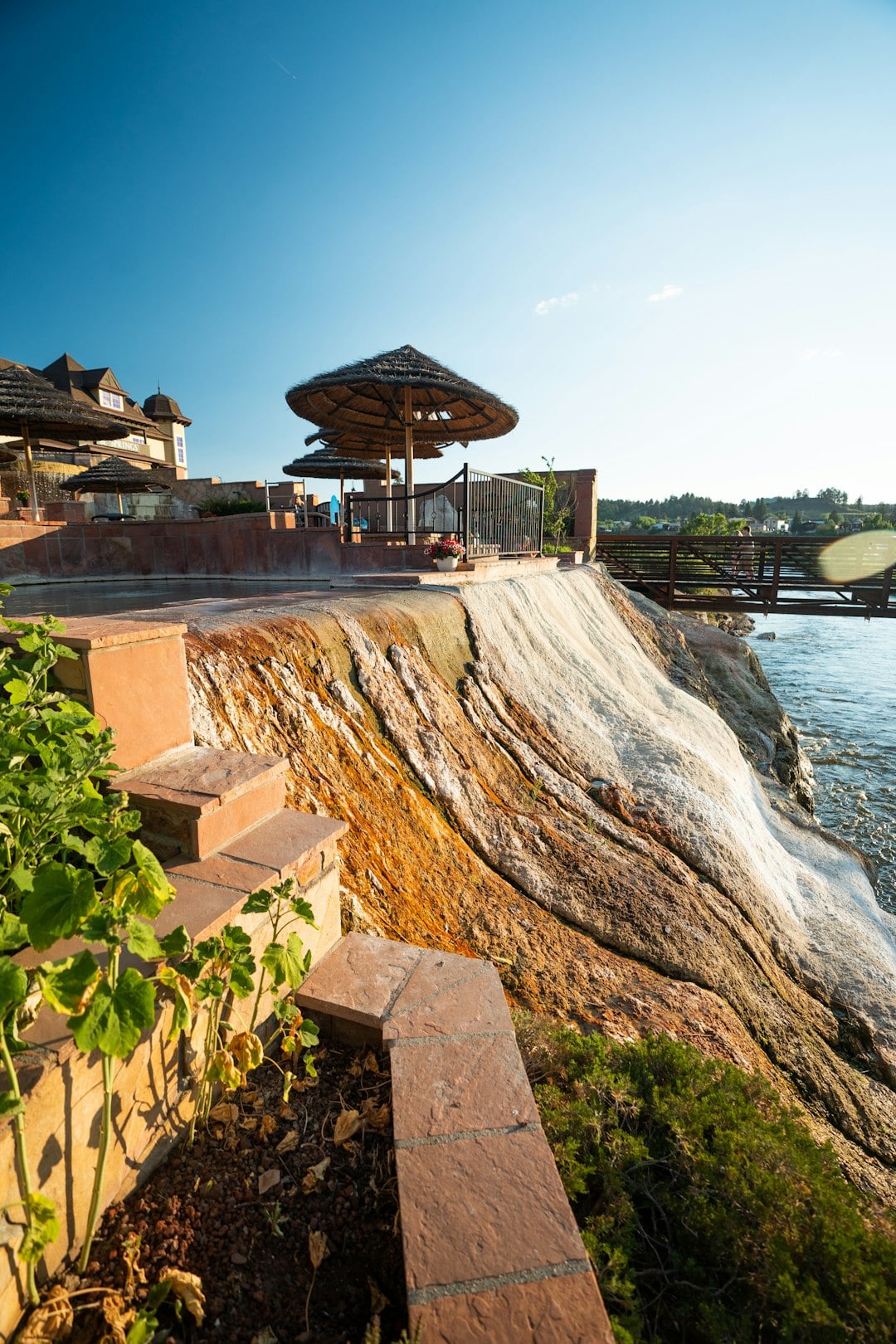 Mineral-stained cliff descends to a river, bordered by brick terraces with straw umbrellas and a building. A distant bridge spans the water under a clear sky.