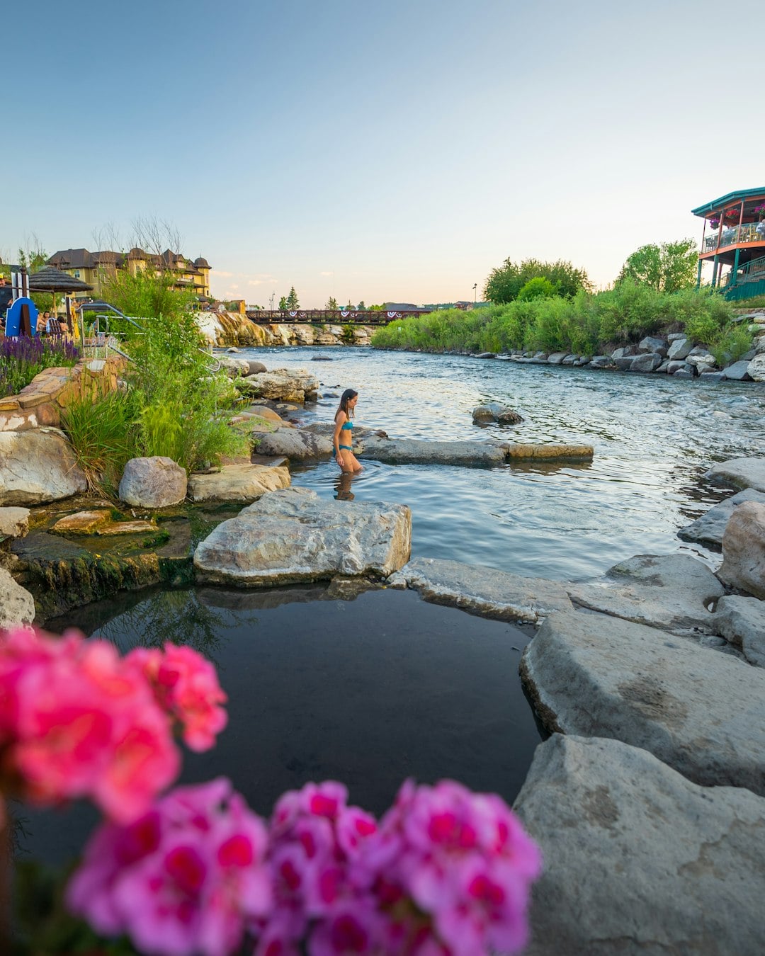 A woman in a blue bikini wades in a rock-lined pool next to a river, with lush banks, a distant bridge, and buildings, framed by pink flowers.