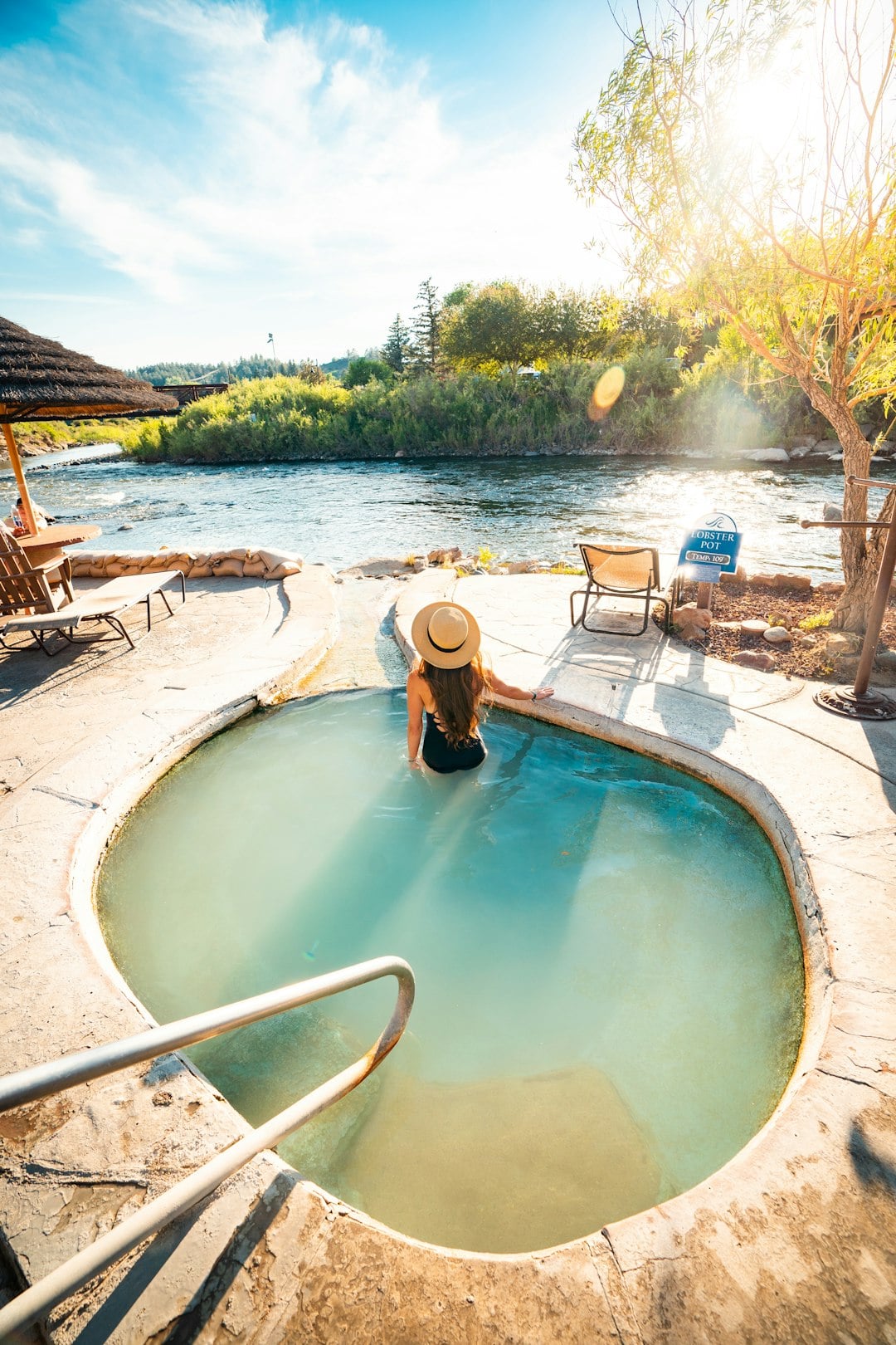 Woman in a straw hat and black swimsuit relaxes in a circular hot spring pool next to a river with green banks. Text: LOBSTER POT TEMP 109°