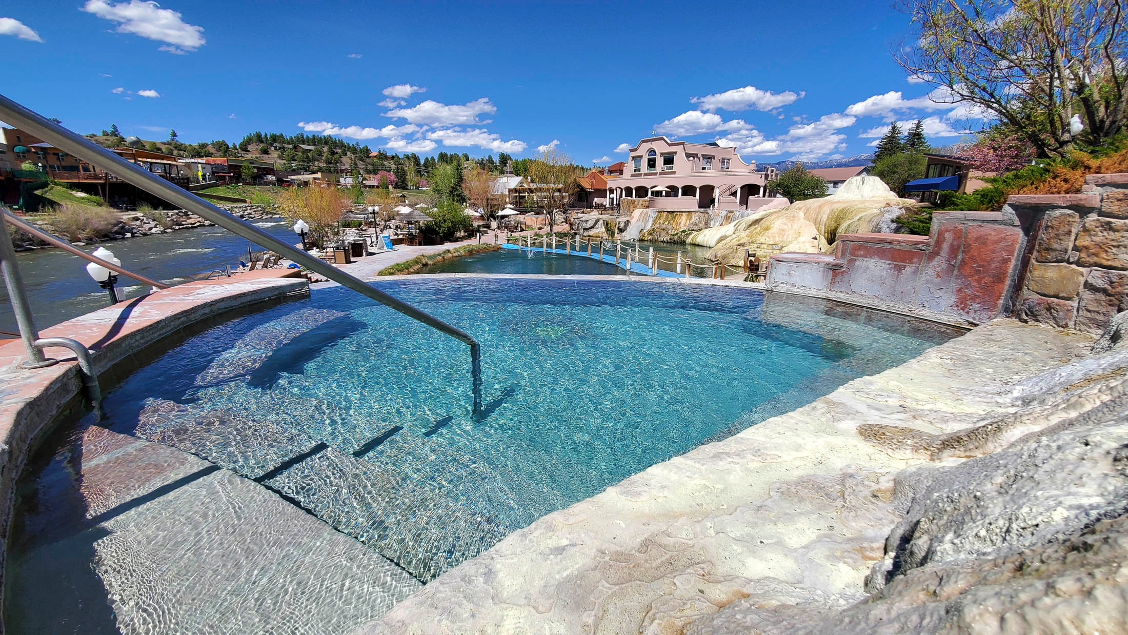 A clear blue hot spring pool with steps and a handrail sits by a river, alongside other pools, a large pink building, and hills under a bright blue sky.