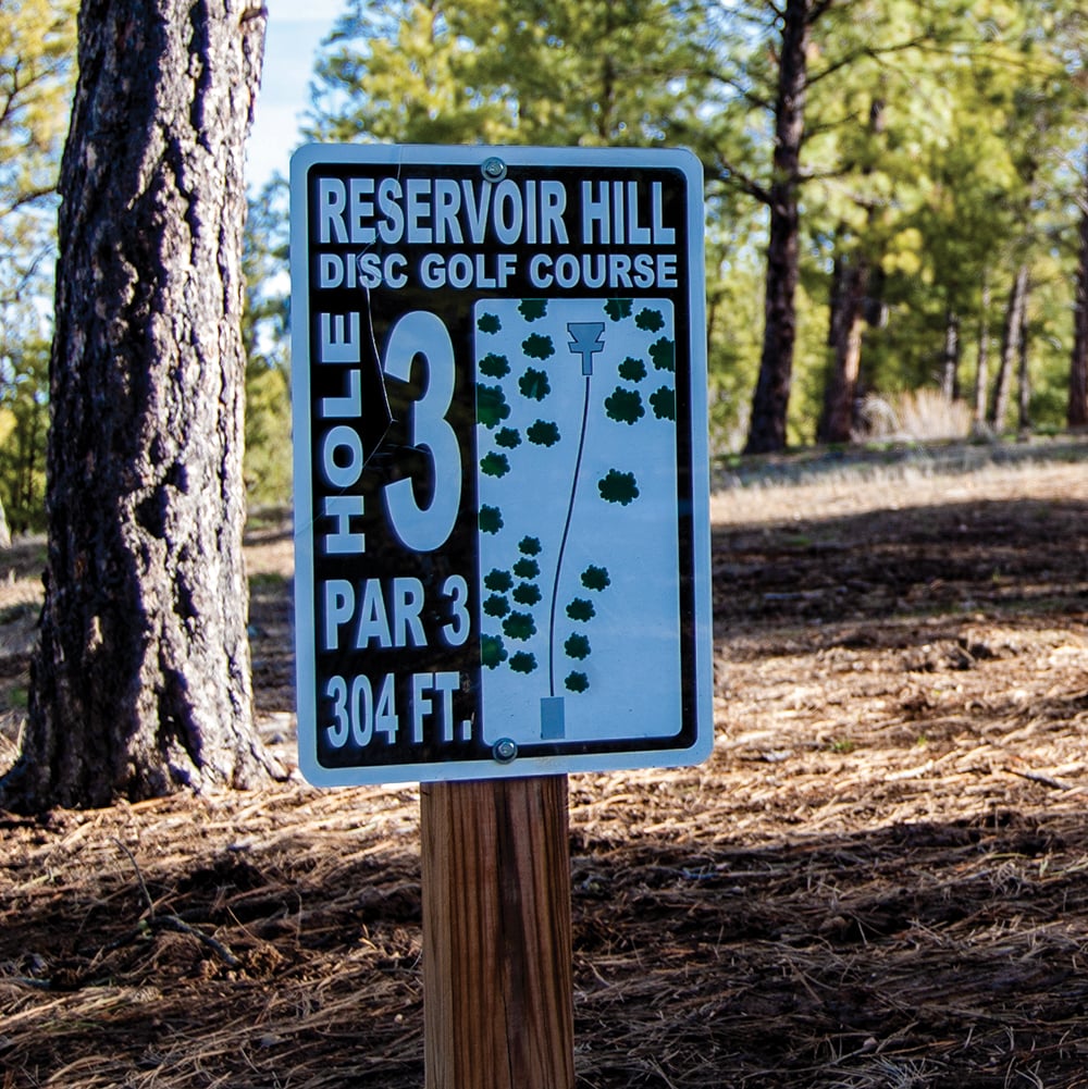 A disc golf course sign stands on a wooden post, displaying hole information and a map in a sunny, forested area. Text: RESERVOIR HILL DISC GOLF COURSE HOLE 3 PAR 3 304 FT.