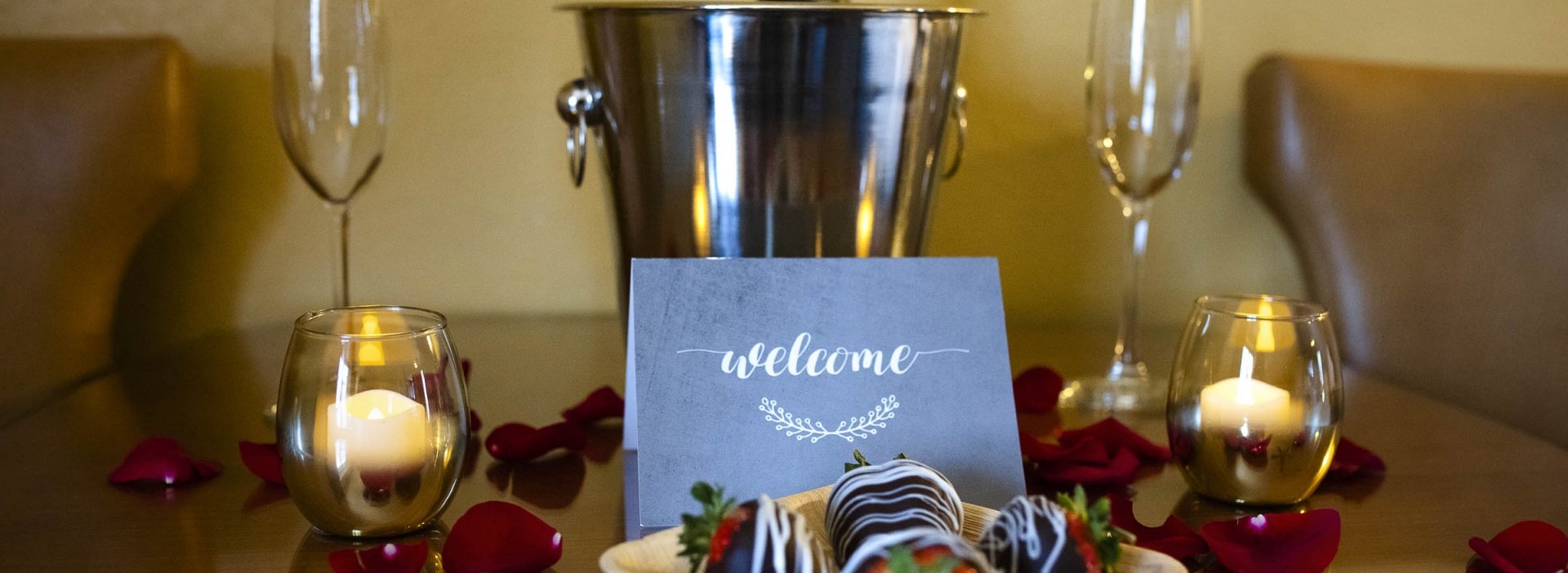 Chocolate-covered strawberries, champagne in an ice bucket, glasses laid out on a wooden table decorated with rose petals