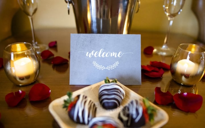 A champagne bottle in an ice bucket, two flutes, and a plate of chocolate-covered strawberries are arranged on a wooden table. Candles and scattered red rose petals set a romantic mood. A card reads: 