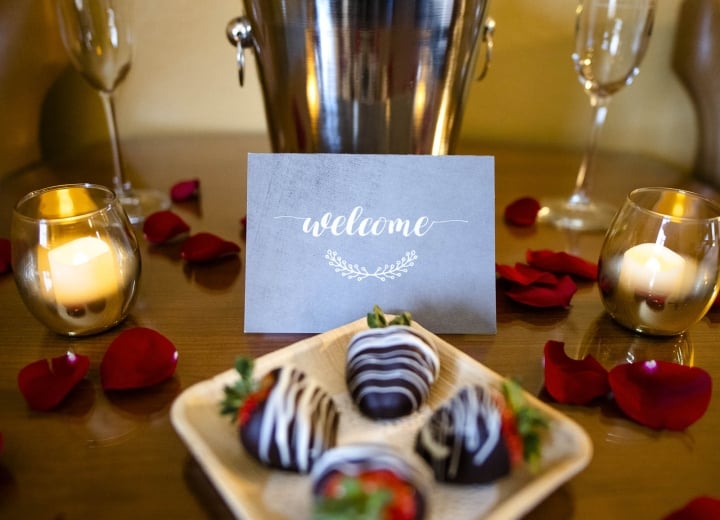 A champagne bottle in an ice bucket, two flutes, and a plate of chocolate-covered strawberries are arranged on a wooden table. Candles and scattered red rose petals set a romantic mood. A card reads: 
