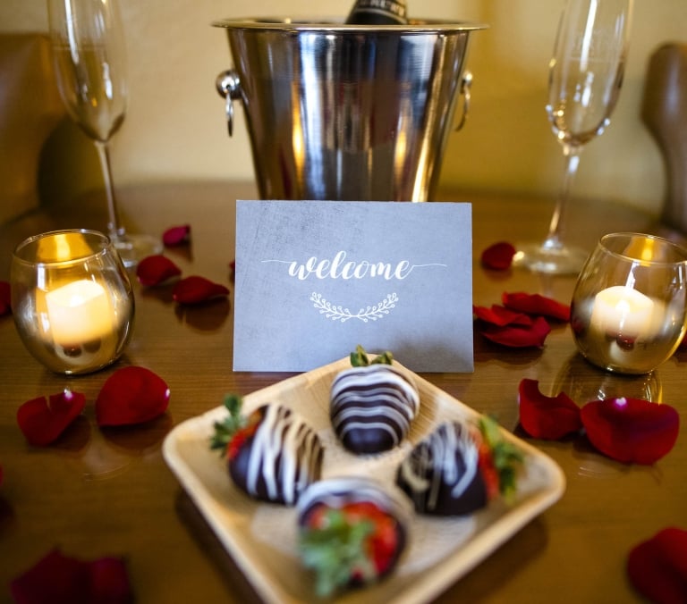 A champagne bottle in an ice bucket, two flutes, and a plate of chocolate-covered strawberries are arranged on a wooden table. Candles and scattered red rose petals set a romantic mood. A card reads: 
