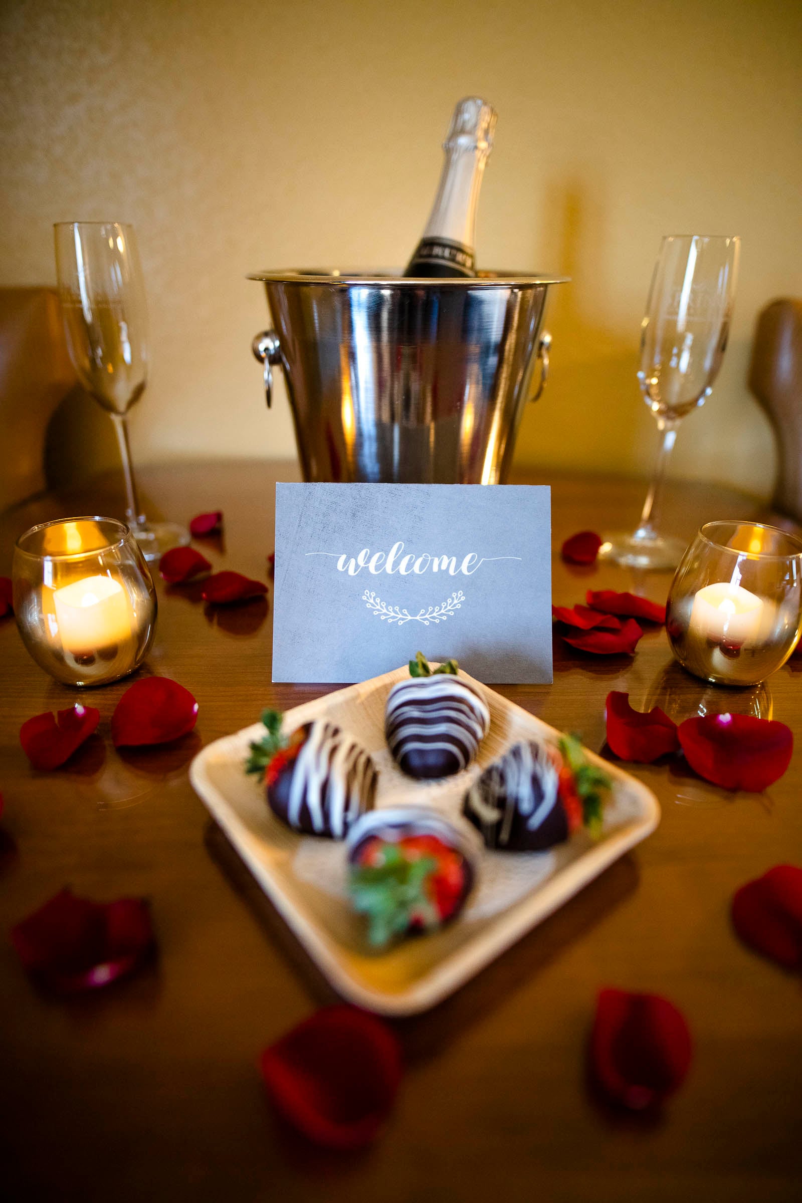 A champagne bottle in an ice bucket, two flutes, and a plate of chocolate-covered strawberries are arranged on a wooden table. Candles and scattered red rose petals set a romantic mood. A card reads: 