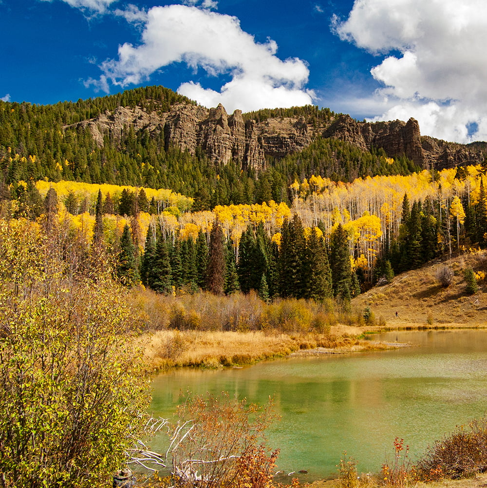 A mountain lake reflects vibrant yellow aspen and dark green evergreen trees, with rugged cliffs under a blue, cloudy sky.