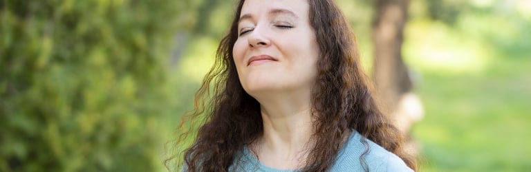 A woman with long curly brown hair smiles with closed eyes, head tilted up, wearing a blue top, outdoors amidst blurred green foliage and trees.