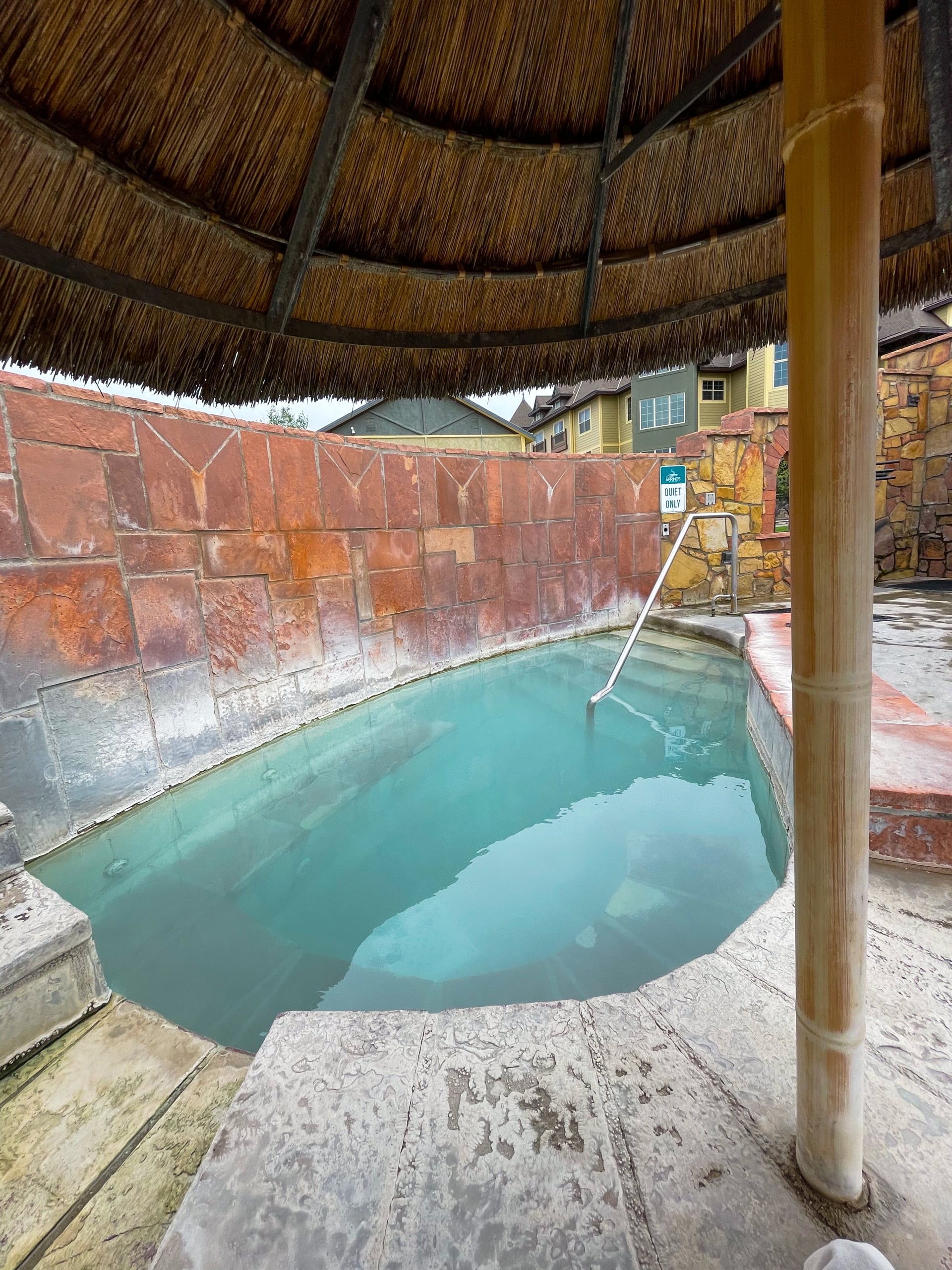 A light blue hot spring pool with a metal handrail sits under a thatched roof, surrounded by red stone walls and distant apartment buildings. Sign reads: 