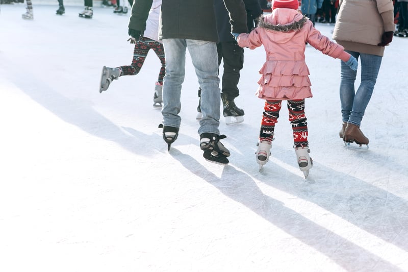 A child in a pink coat and patterned leggings learns to ice skate, holding hands with an adult on a bright outdoor rink among other skaters.