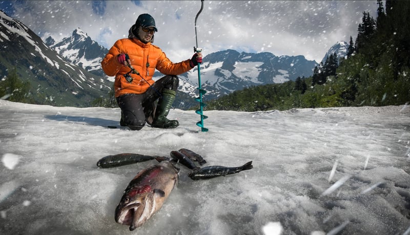 A man in an orange jacket kneels, ice fishing with a rod and auger, several fish caught. Falling snow covers an icy mountain landscape.