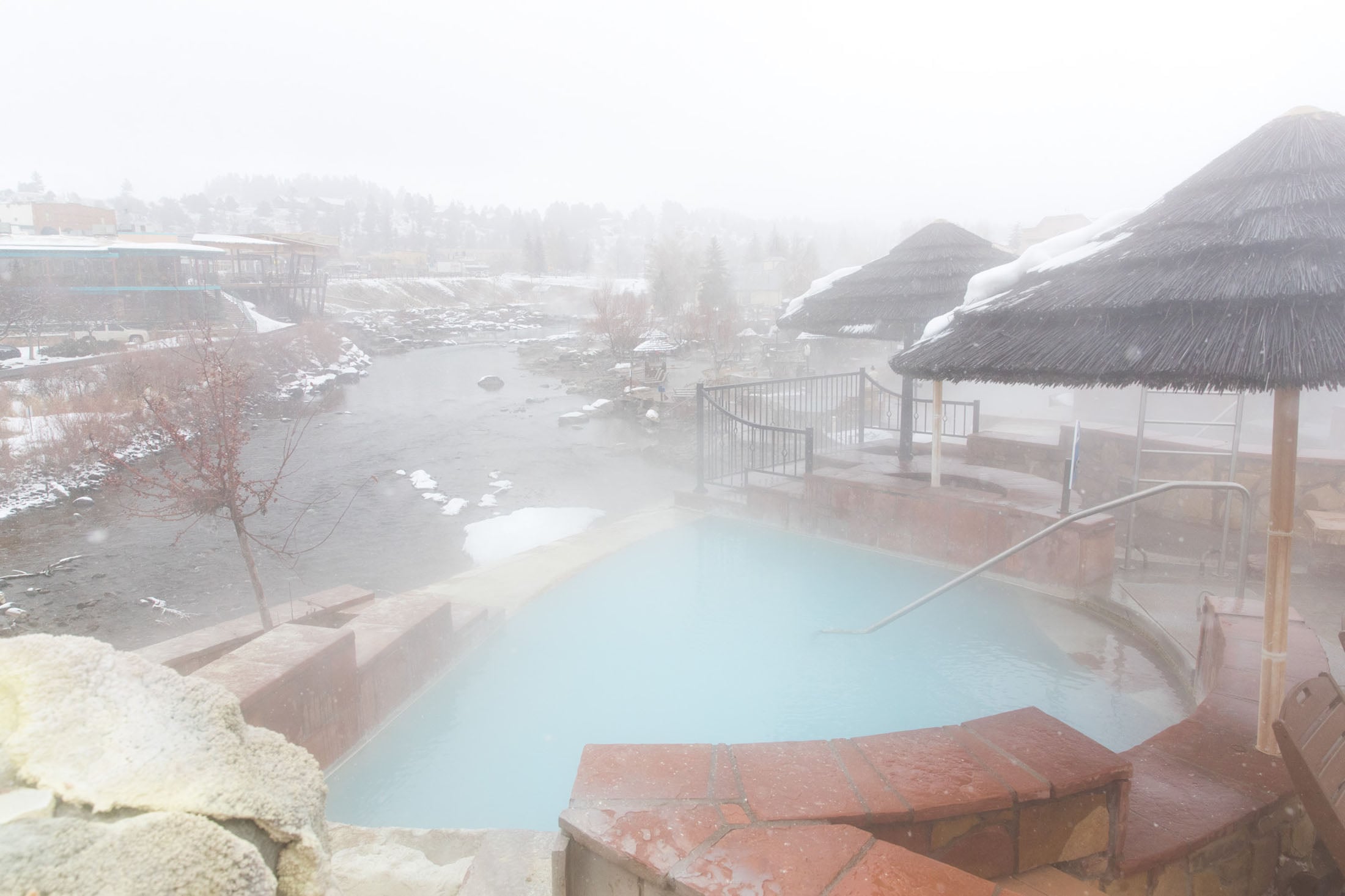 Steaming light blue hot spring pool emits vapor, surrounded by brick coping and snow-dusted thatched umbrellas. It sits beside a snowy river, with a misty town in the distance.