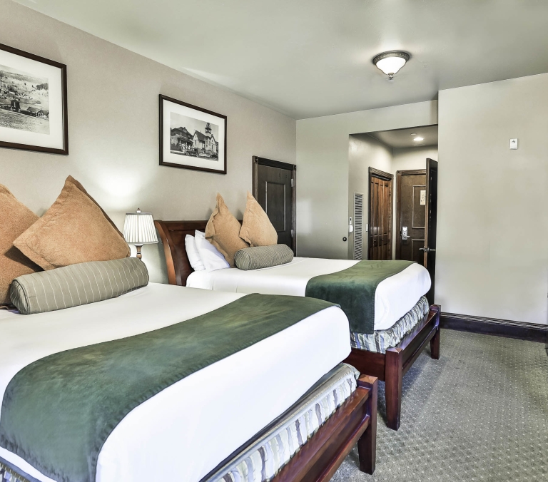 Two queen beds with white linens, green throws, and decorative pillows fill a hotel room. Framed artwork hangs on light walls, alongside dark wooden doors and a dresser with a TV.