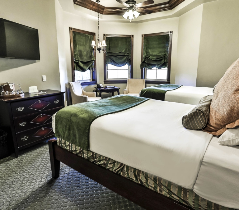 A well-appointed hotel room features two made beds with green throws, a wall-mounted TV, dark dresser, and windows with green velvet blinds under a chandelier and ceiling fan.