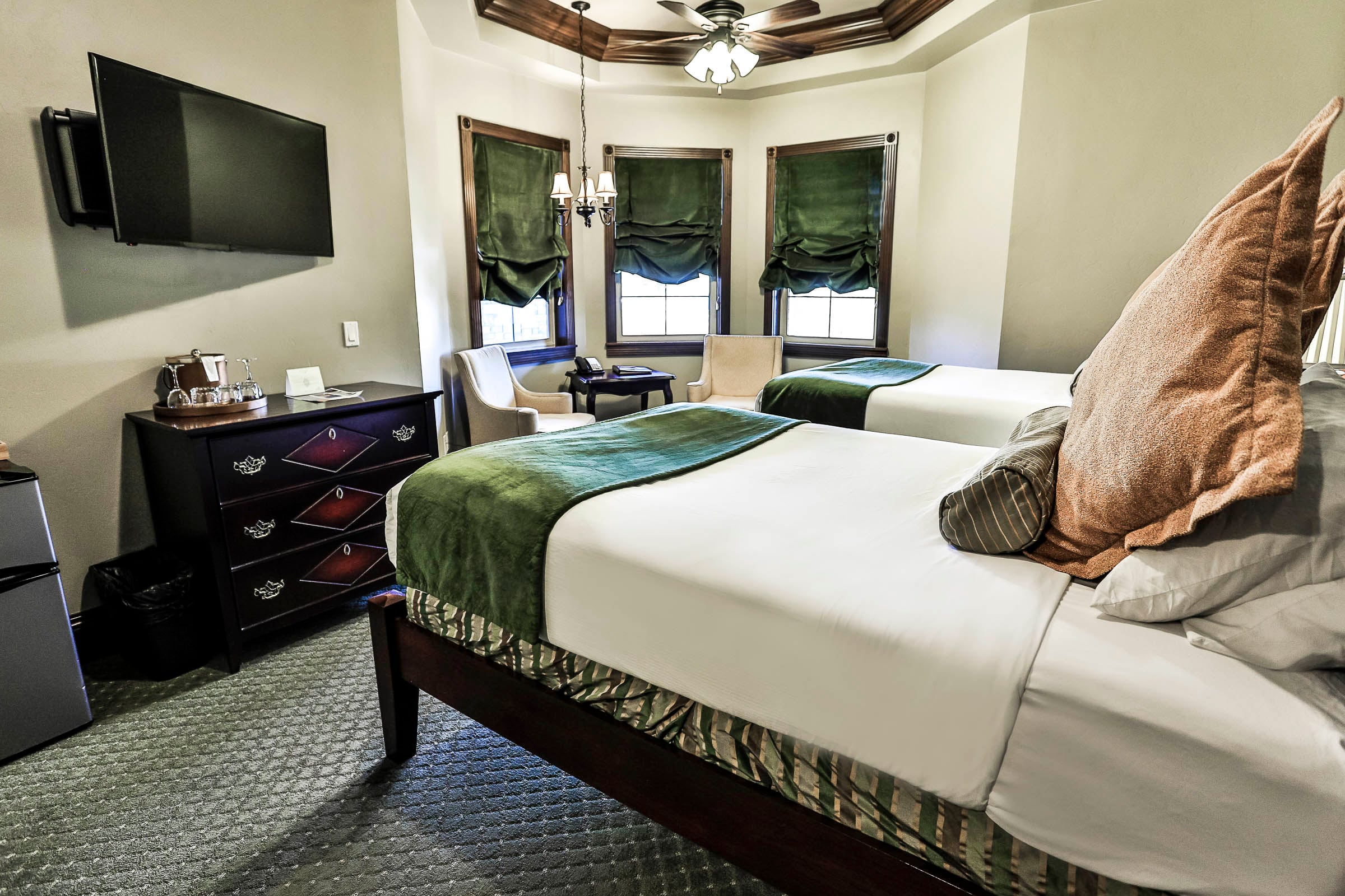 A well-appointed hotel room features two made beds with green throws, a wall-mounted TV, dark dresser, and windows with green velvet blinds under a chandelier and ceiling fan.