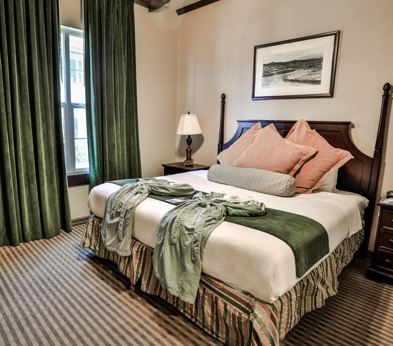 A neatly made bed with a dark wooden headboard, white sheets, a green runner, and two light green robes, in a hotel room with striped carpet and green curtains.