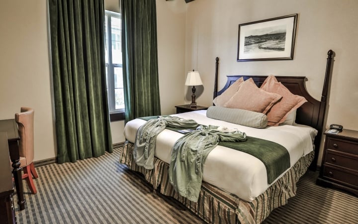 A made bed with a green runner and robe sits in a hotel room. It features dark green curtains, a striped carpet, and a framed landscape picture on the wall.