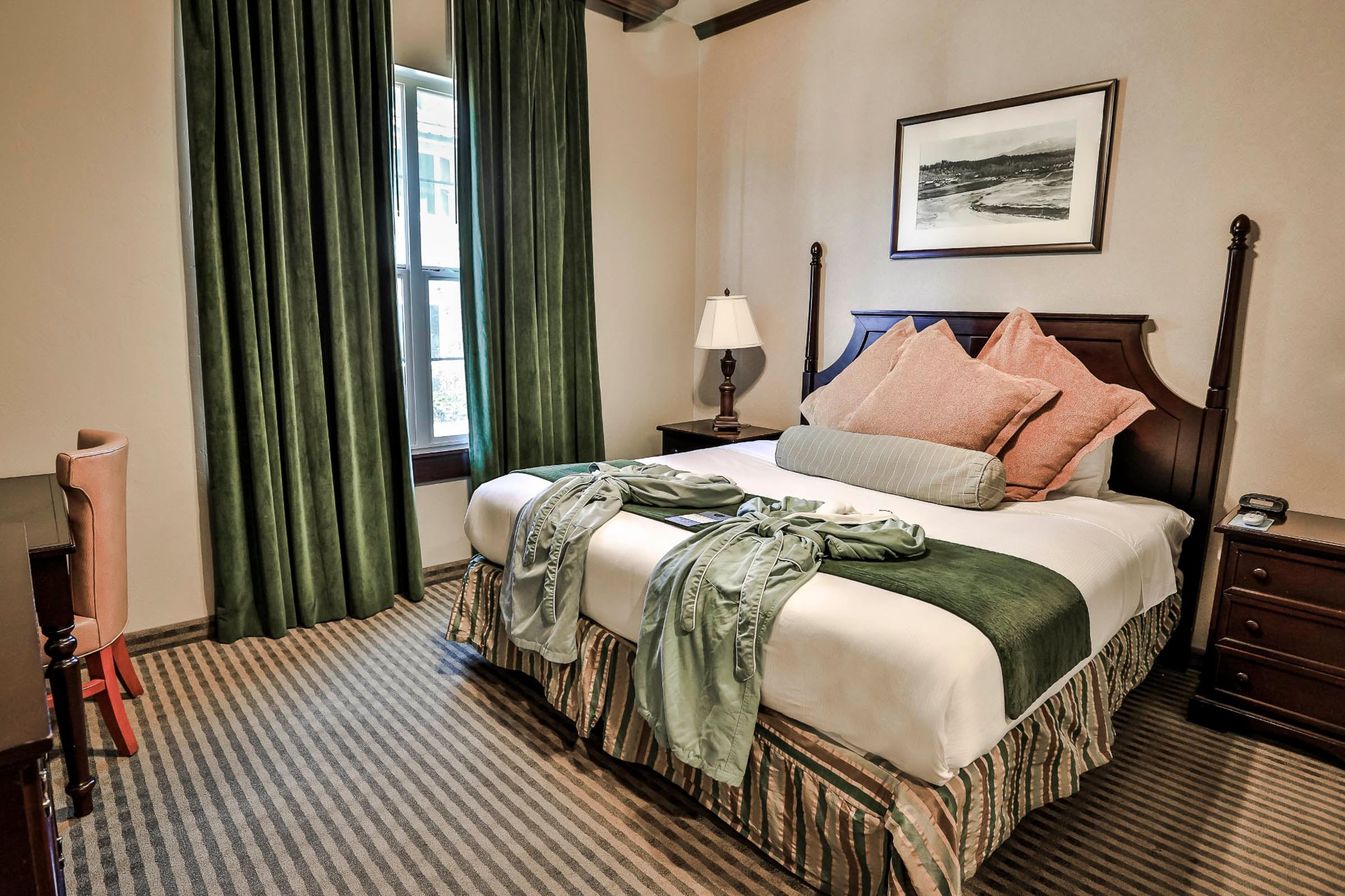 A made bed with a green runner and robe sits in a hotel room. It features dark green curtains, a striped carpet, and a framed landscape picture on the wall.