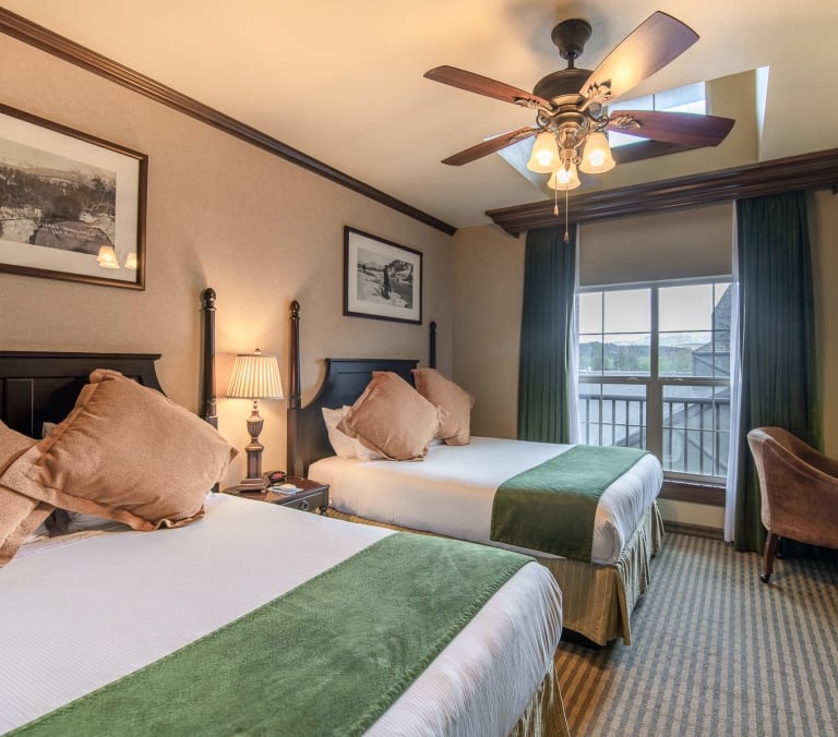 Two queen beds with green throws furnish a hotel room featuring a window, ceiling fan, desk, armchair, and framed artwork 